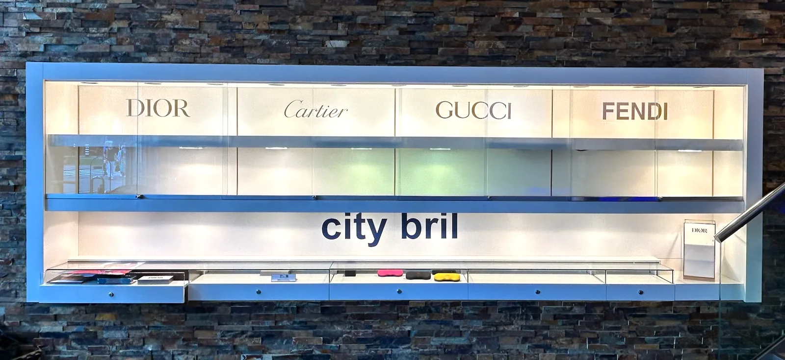 Na: witte vitrinewand met de merknamen Dior, Cartier, Gucci, Fendi en city bril
